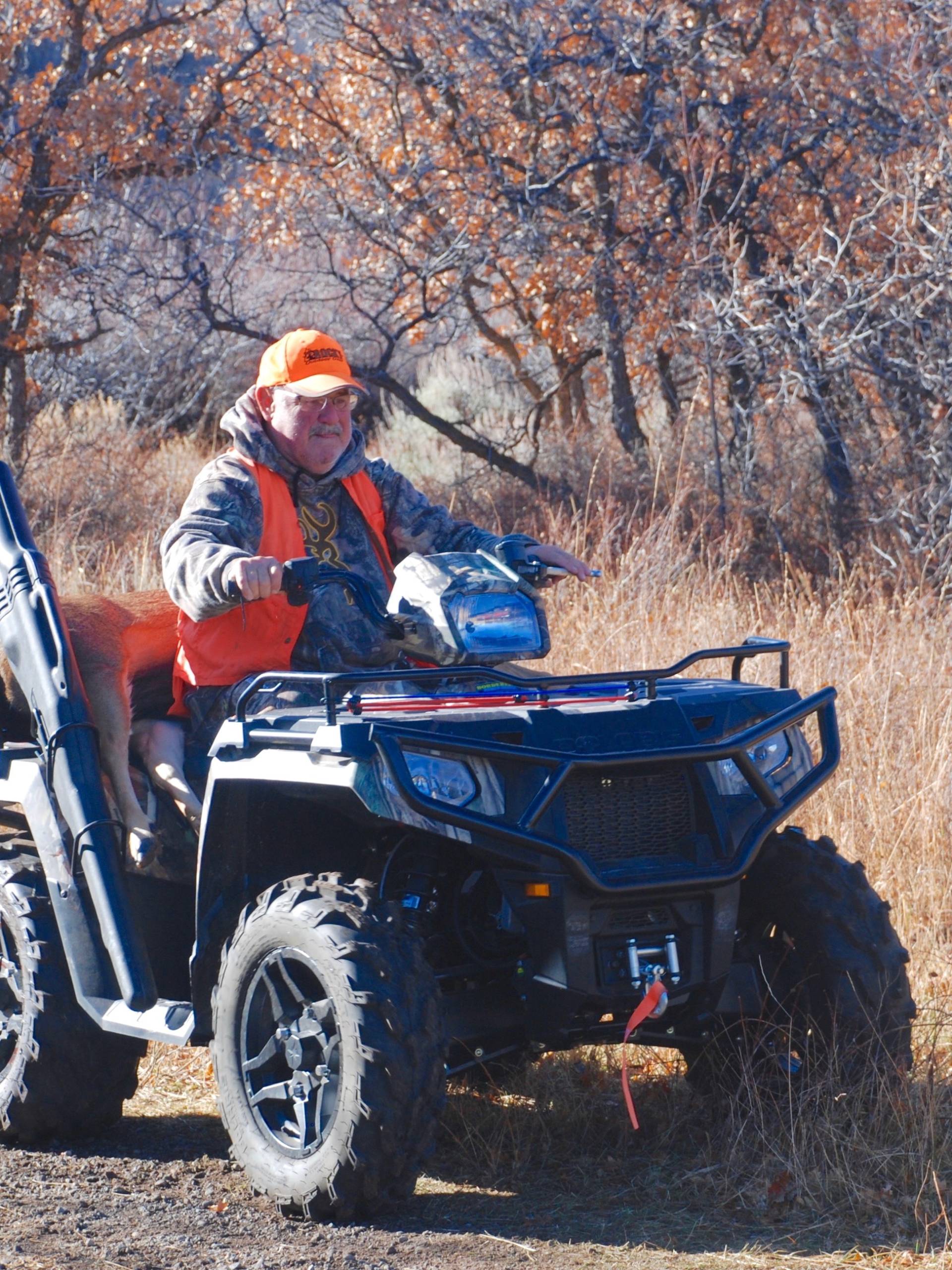 A hunter is sitting on a quad with his firearm by his side. He is transporting his freshly hunted deer.