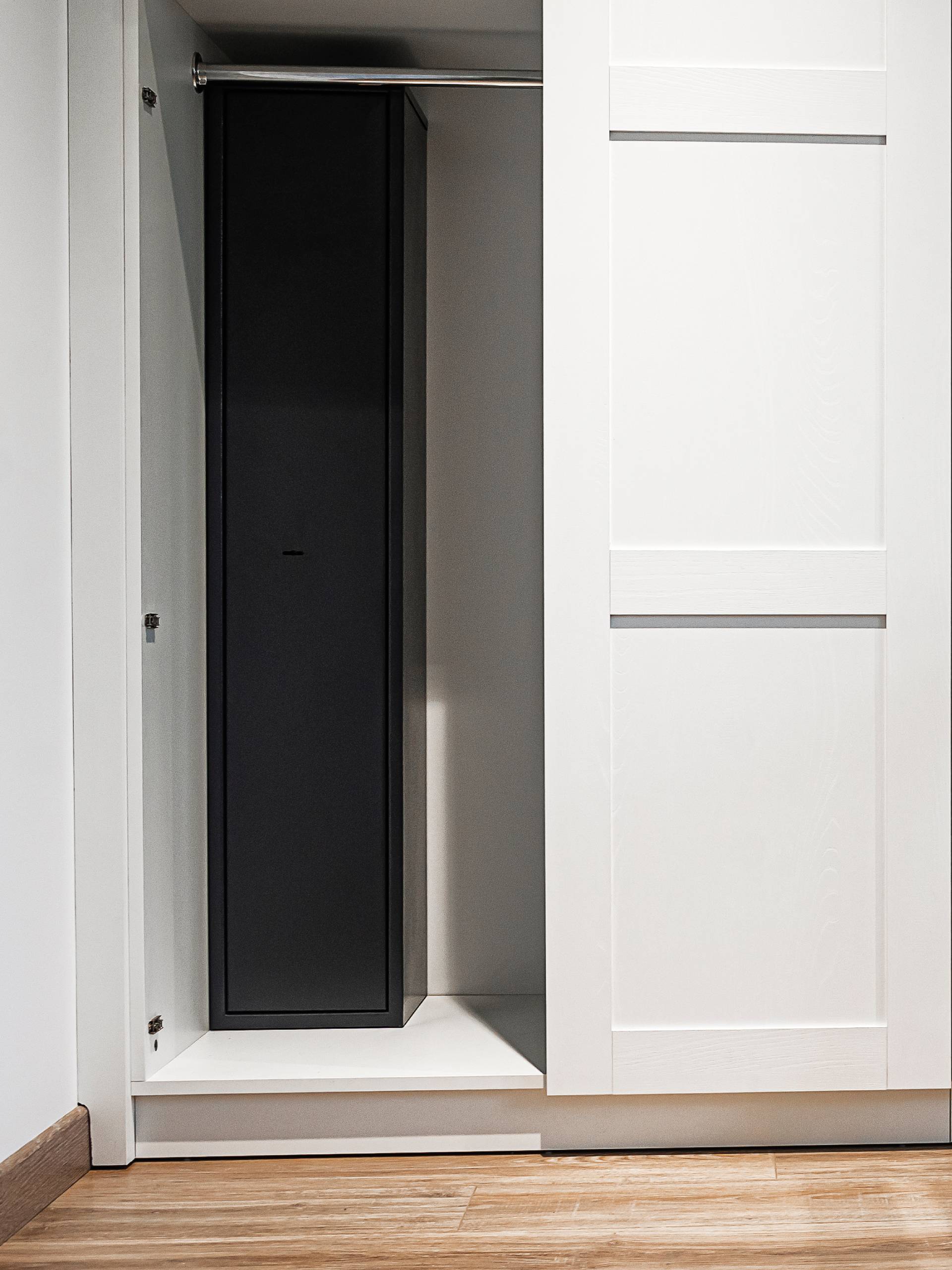 A tall, slim gun safe mounted snugly next to the wall inside a wardrobe with an empty shelf and closet rod.