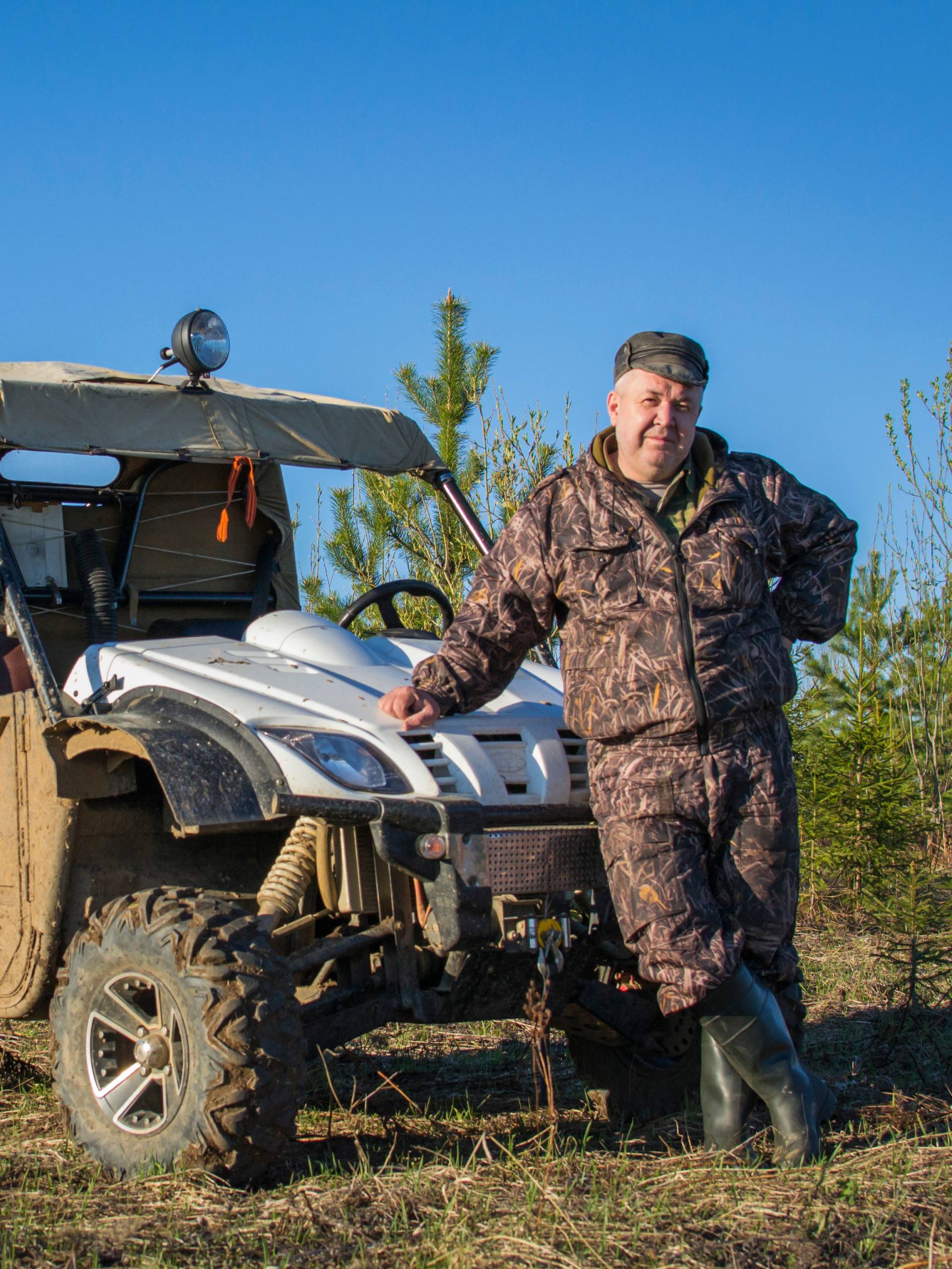 Man wearing camouflage gear standing in front of a UTV covered in mud in a large field outdoors on a sunny day.