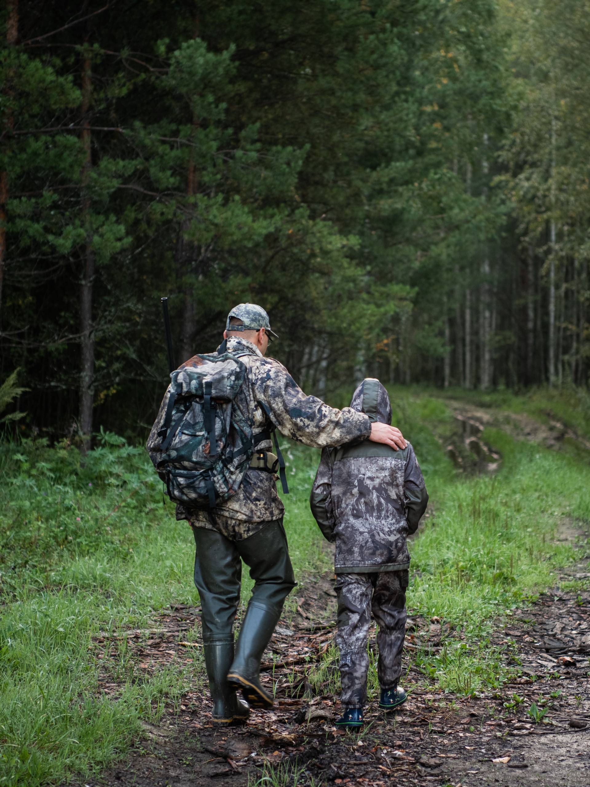 An older hunter resting his hand on his child’s shoulder as the pair walk along a muddy path into the woods.