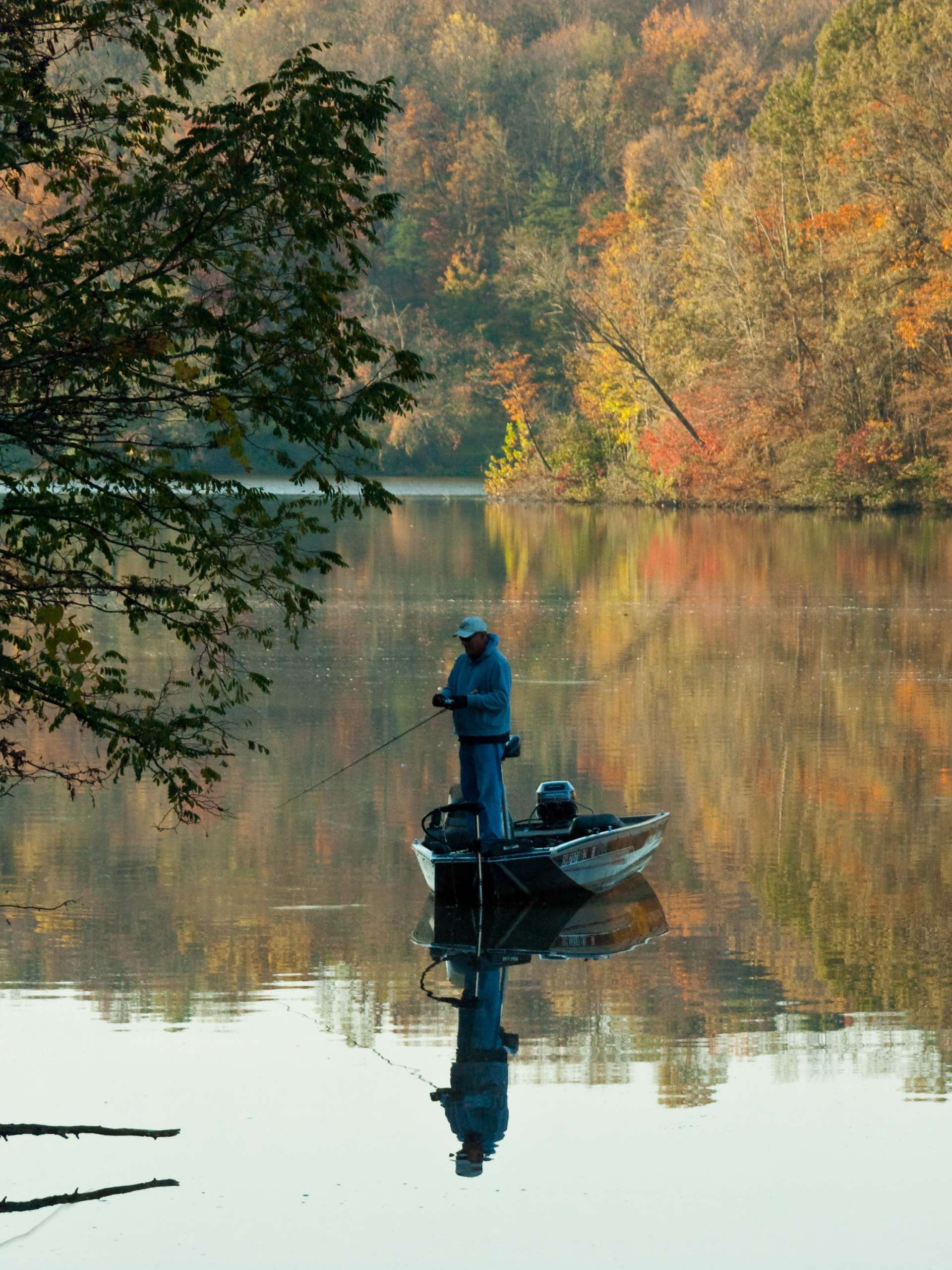 A fishing enthusiast stands on a boat in the middle of a lake fishing. The leaves on the trees are yellow.