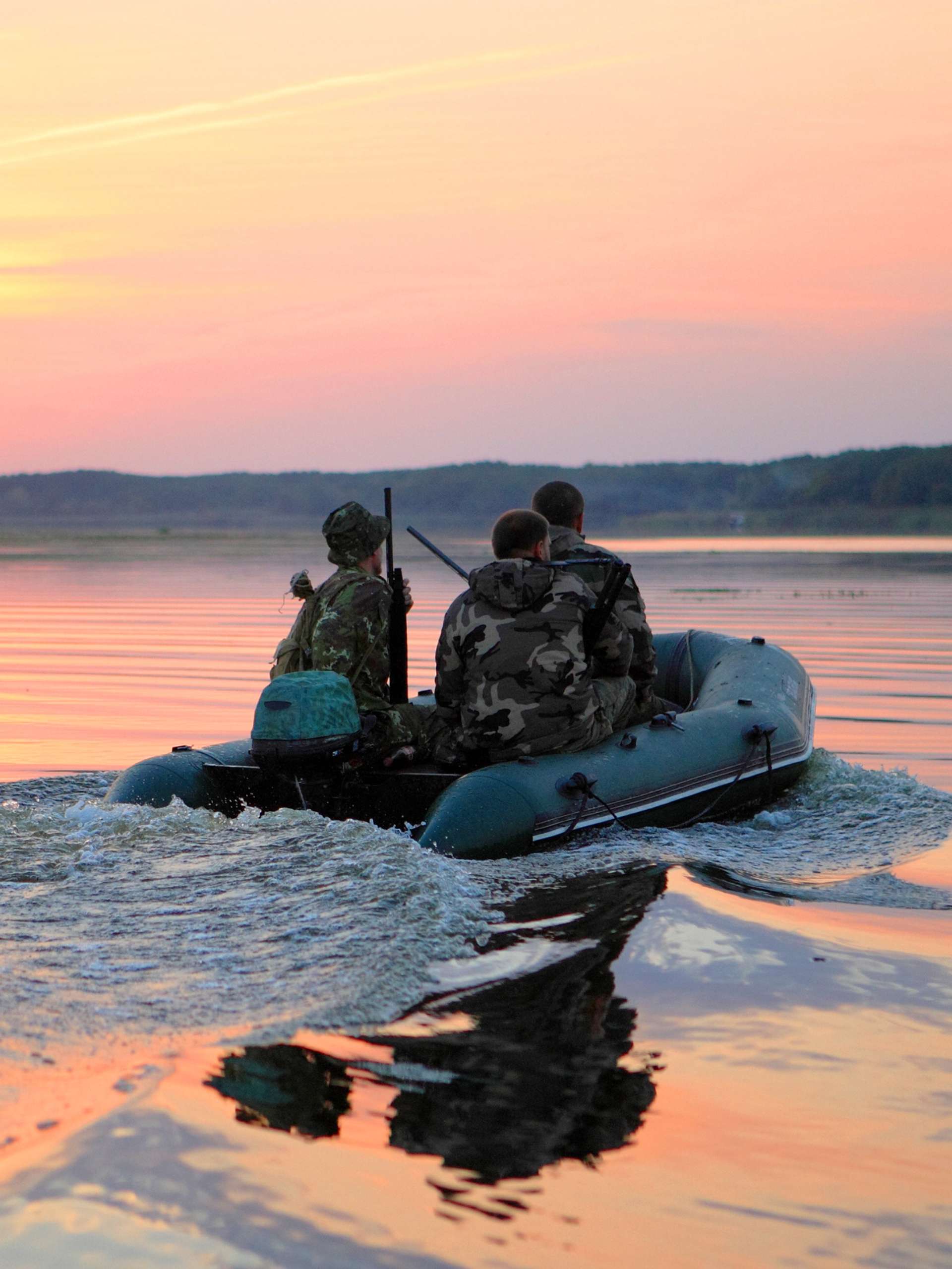 Three people in camouflage hunting gear holding their firearms as they ride a motorboat down a wide river.