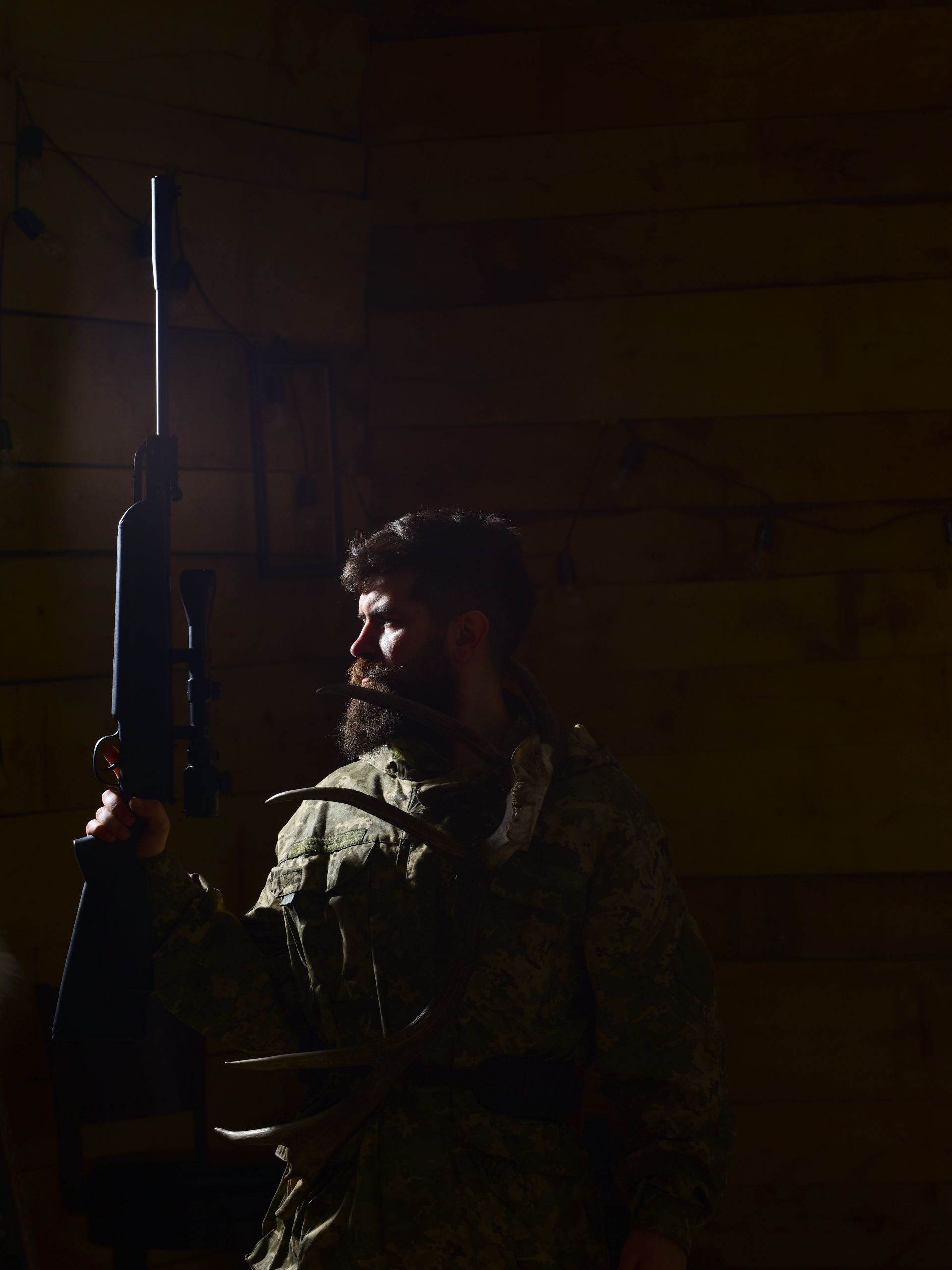 A man stands in the dark wearing camouflaged hunting gear. He holds a rifle in his hand as he stares into the distance.