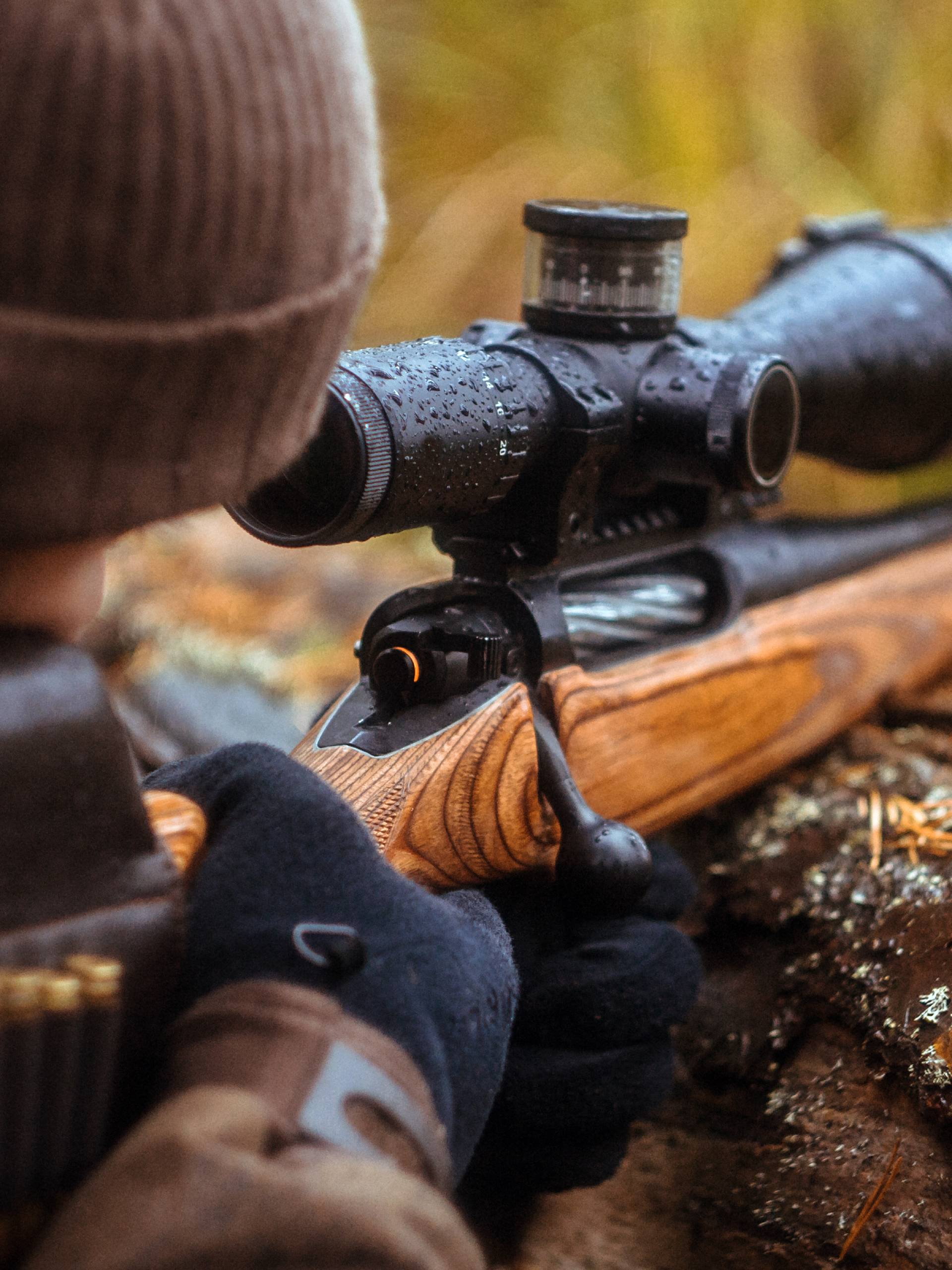 A person dressed in a brown coat and hat rests a rifle with a telescopic sight on it on top of a log.