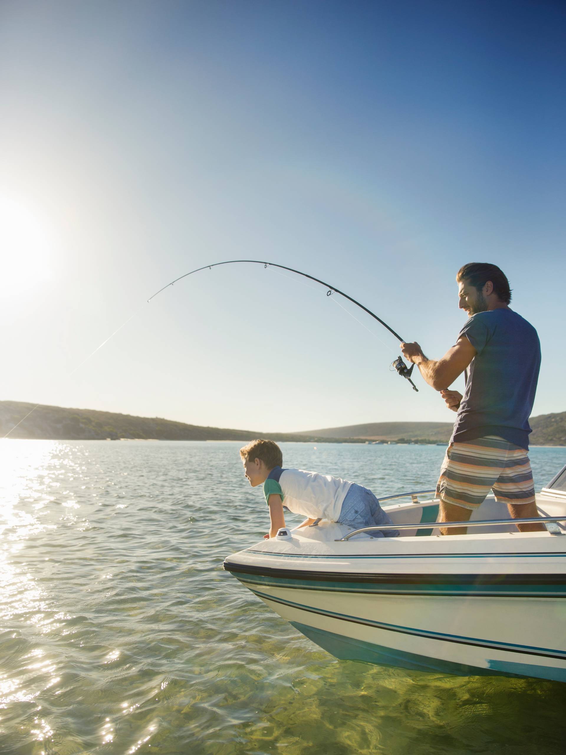 A young boy looks over the side of a white, still boat as an adult male stands next to the boy, holding a fishing pole.