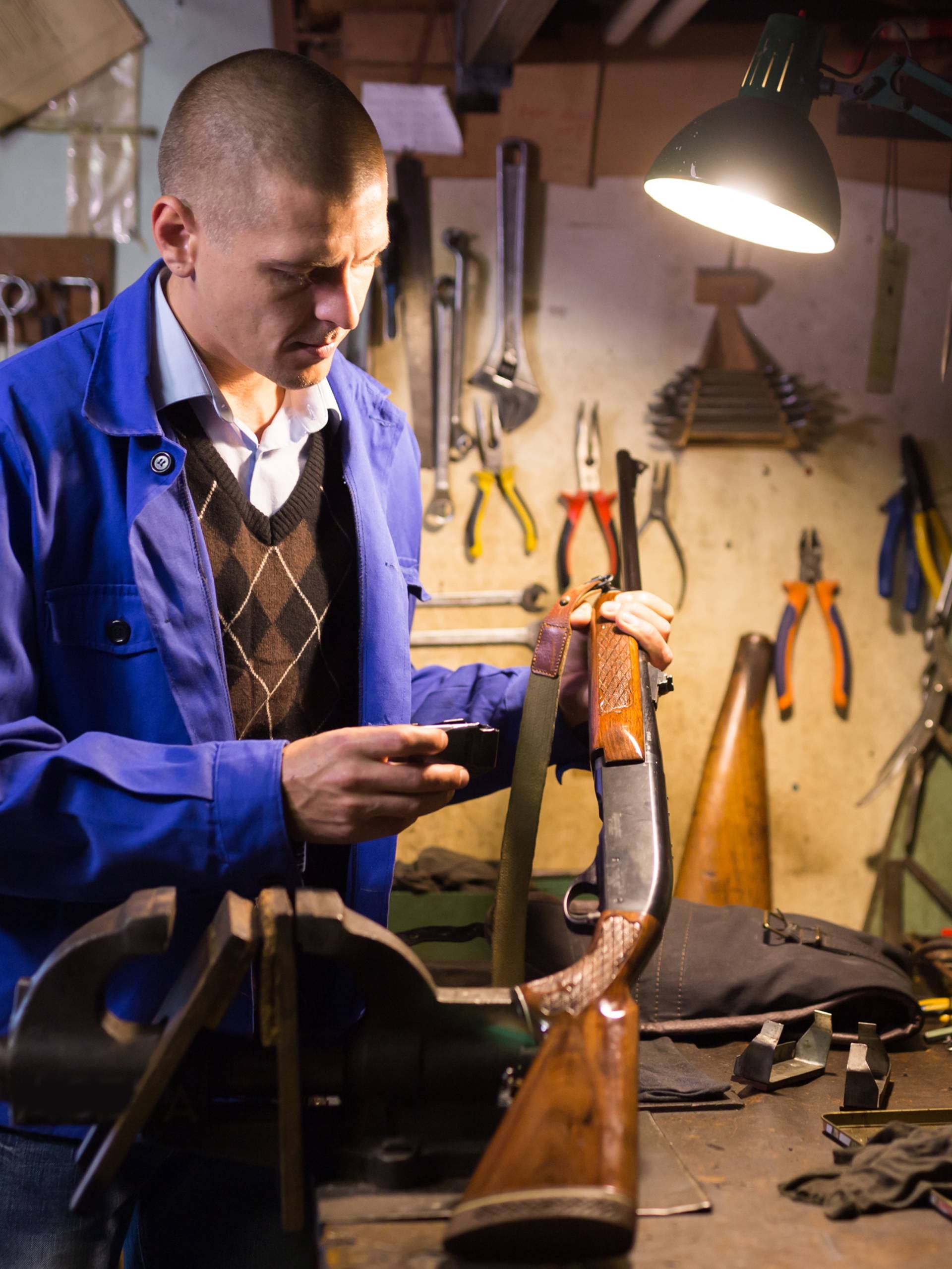 A man in a blue jacket works at a cluttered workshop with tools on the walls, a rifle, a vise, and a focused overhead lamp.