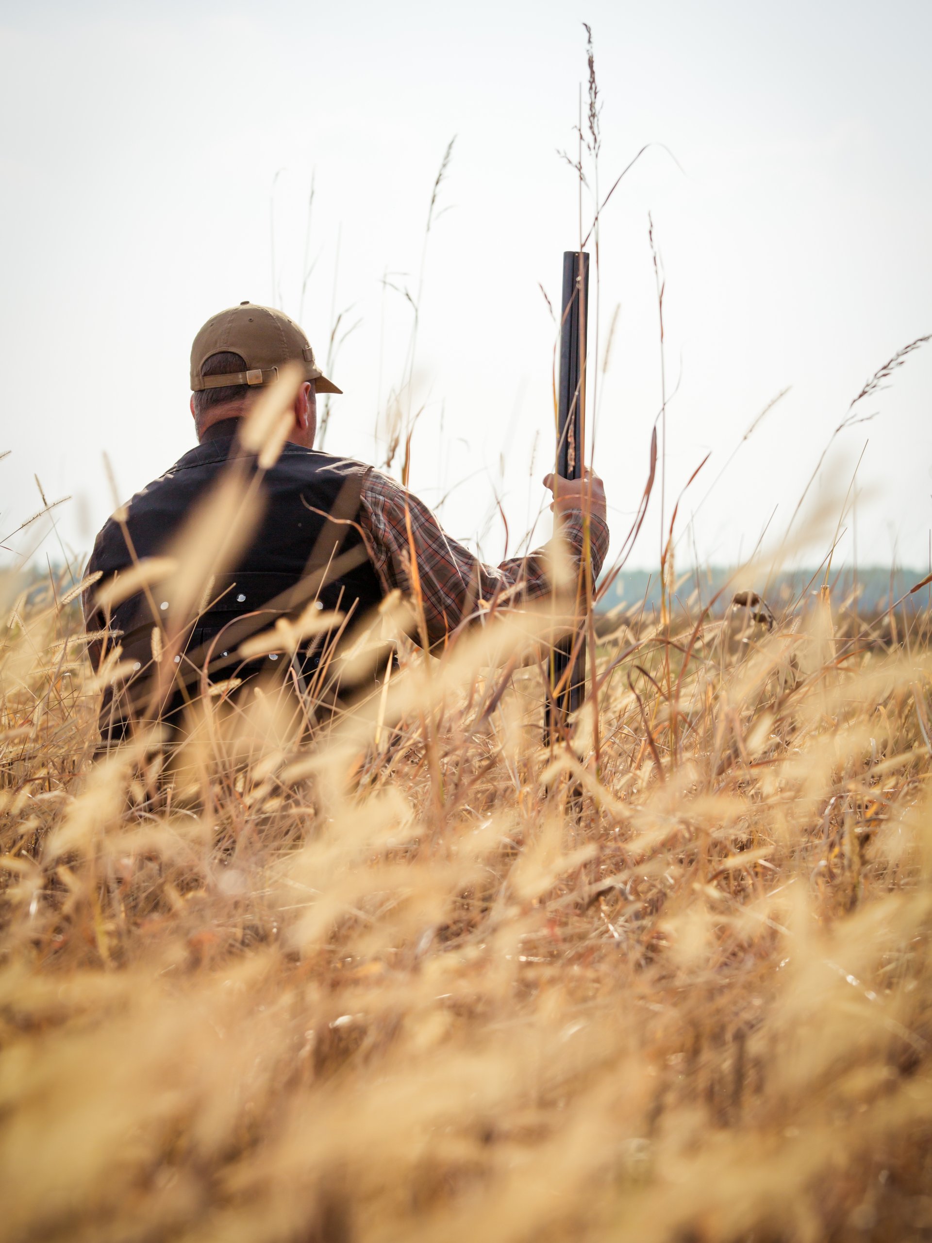 A person crouches in a field full of tall, tan foliage, holding a large gun. The sky around them is gray.