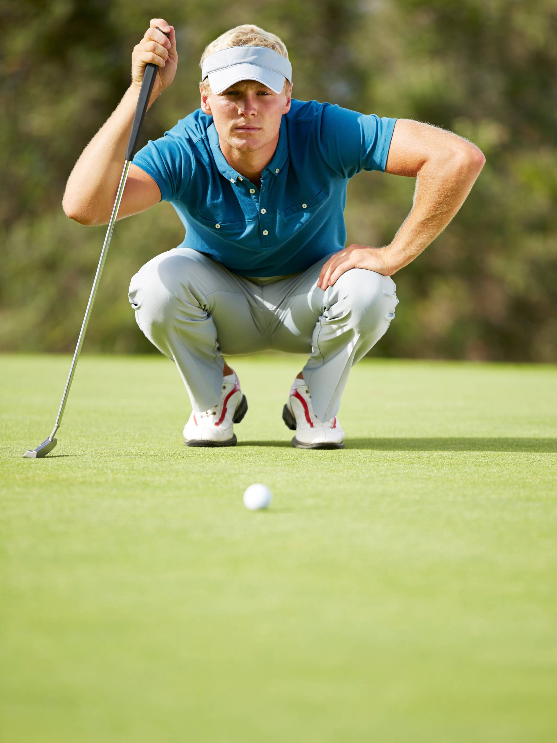 A man wearing a teal polo shirt, cream-colored pants, and a white visor crouches on the putting green at a golf course.