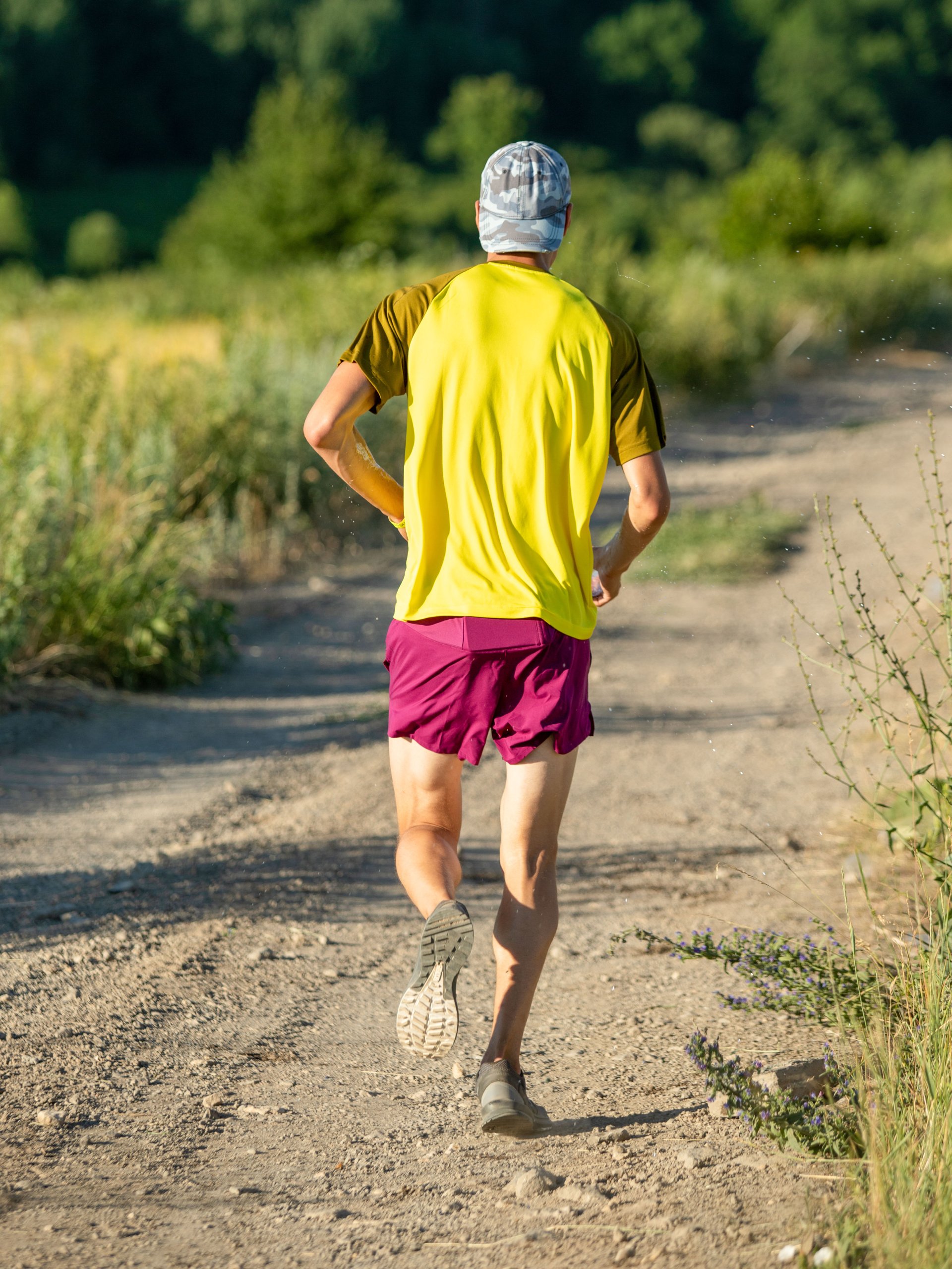 A man wearing a yellow shirt, burgundy shorts, and a white backward baseball cap runs on a dirt path.