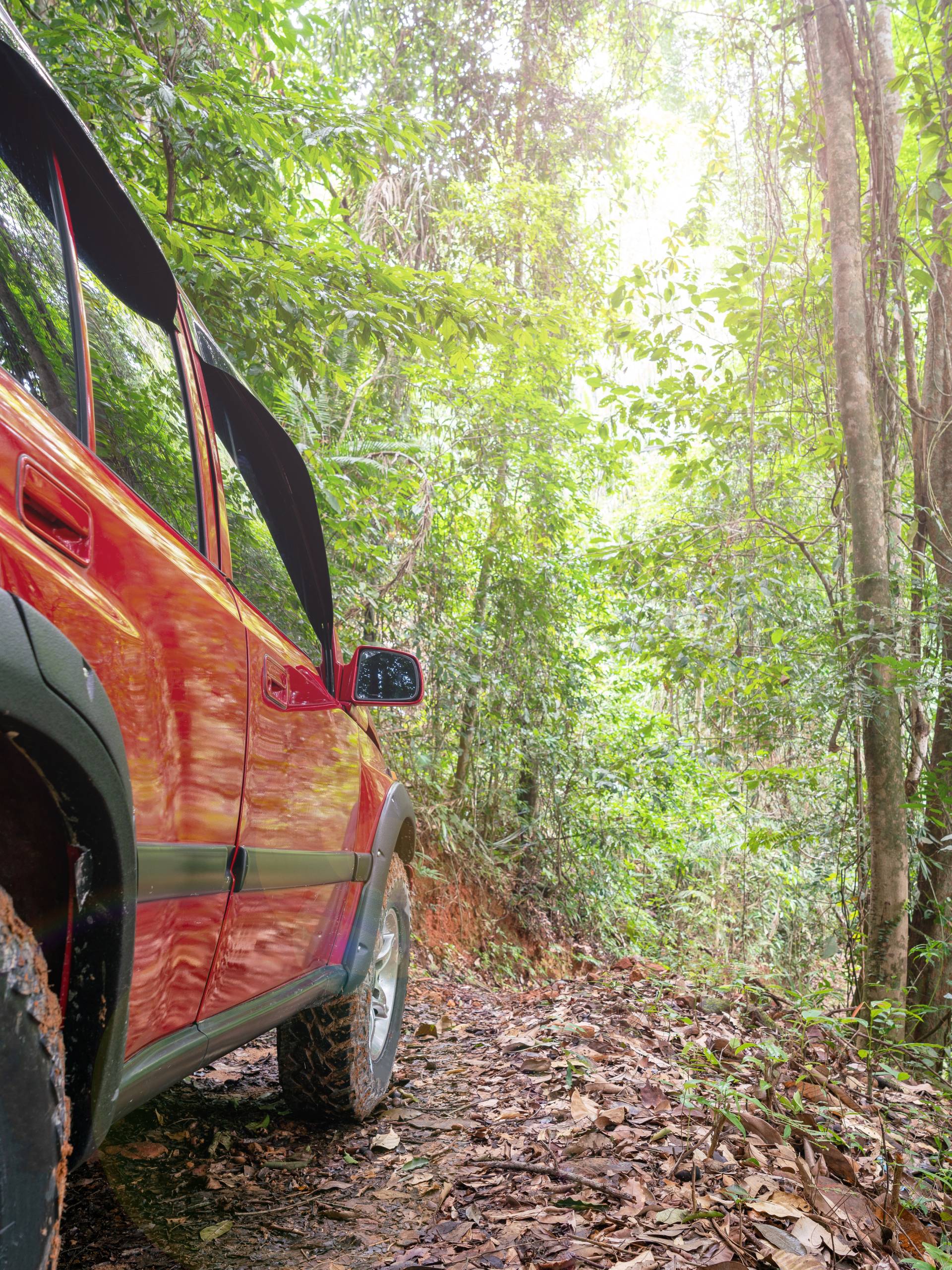 A bright red SUV with muddy tires traversing a thin, leafy trail. The vehicle is driving in a forested area.