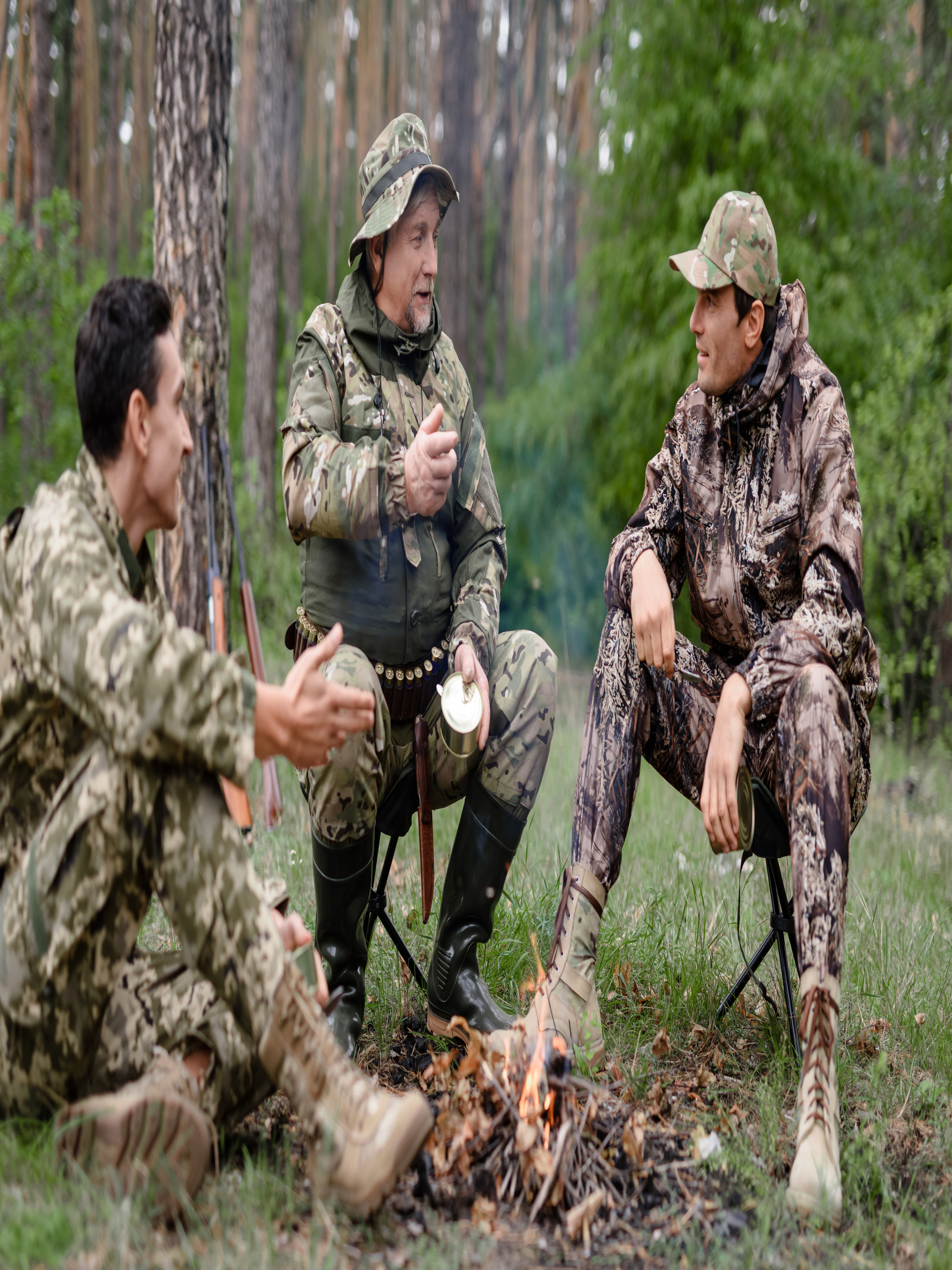 Three men in camouflage gear sitting around a small fire in the forest, talking and taking a break from hunting.