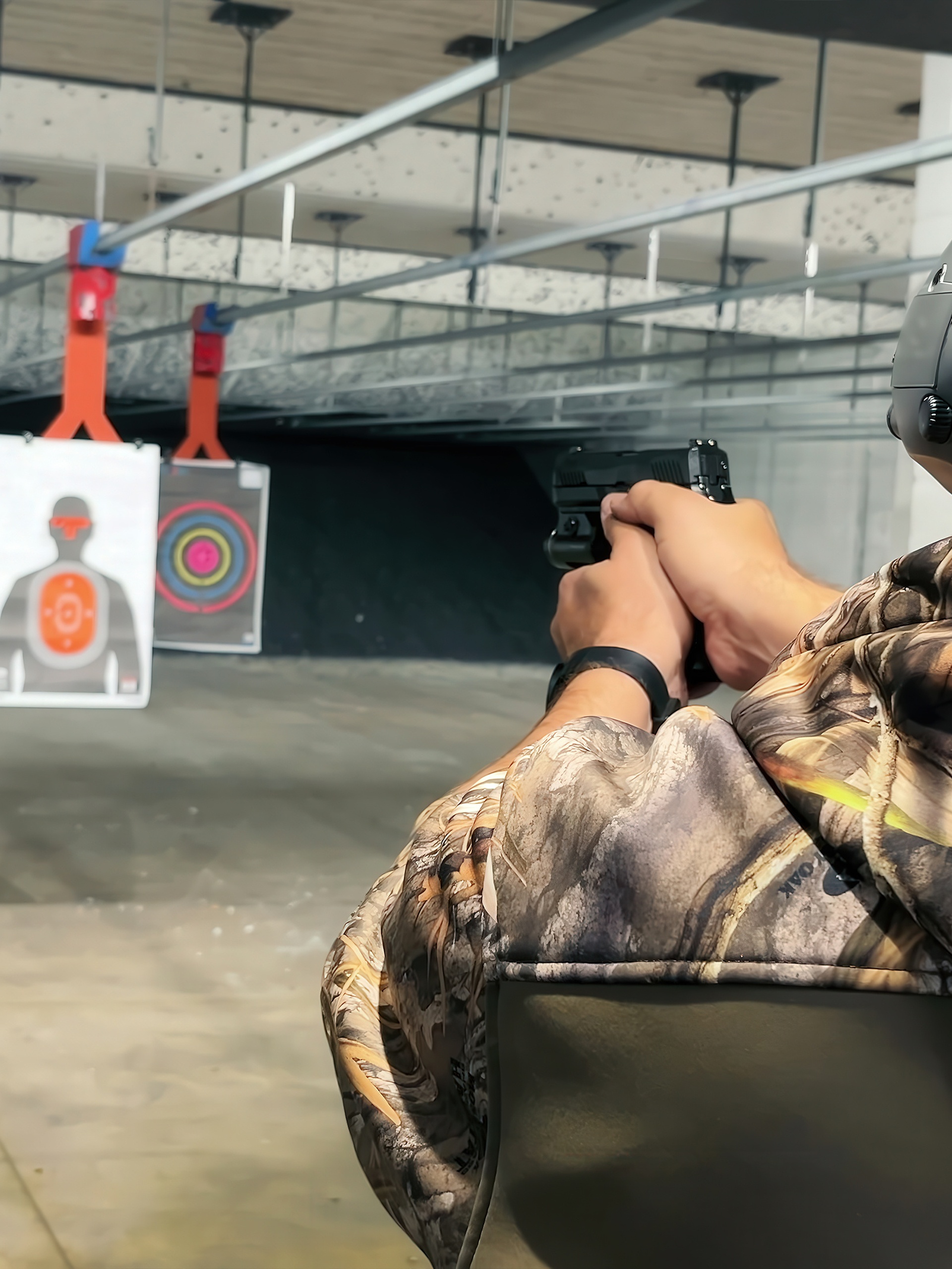 A person wearing earmuffs and camouflage clothing holding a handgun while aiming at targets in an indoor shooting range.
