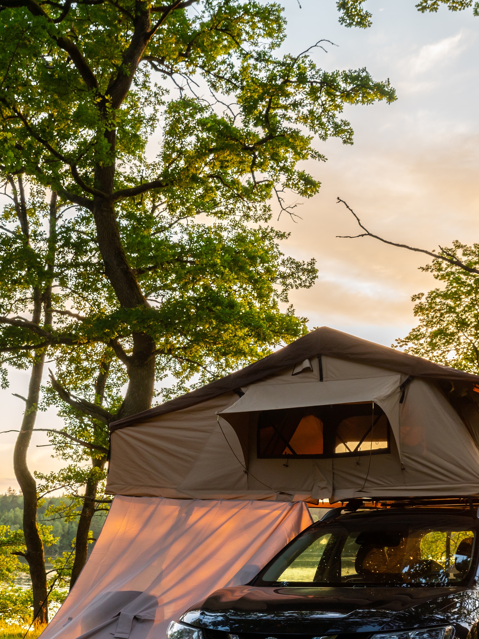 A tan rooftop tent sits on top of a black vehicle that is parked in a wooded area by a body of water.