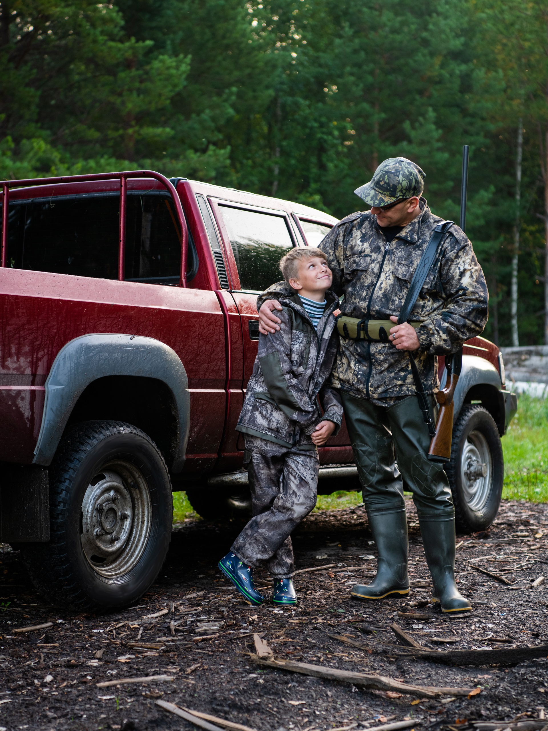 A father and his son standing in front of a red pickup truck in a forest. They are both wearing camo clothes.