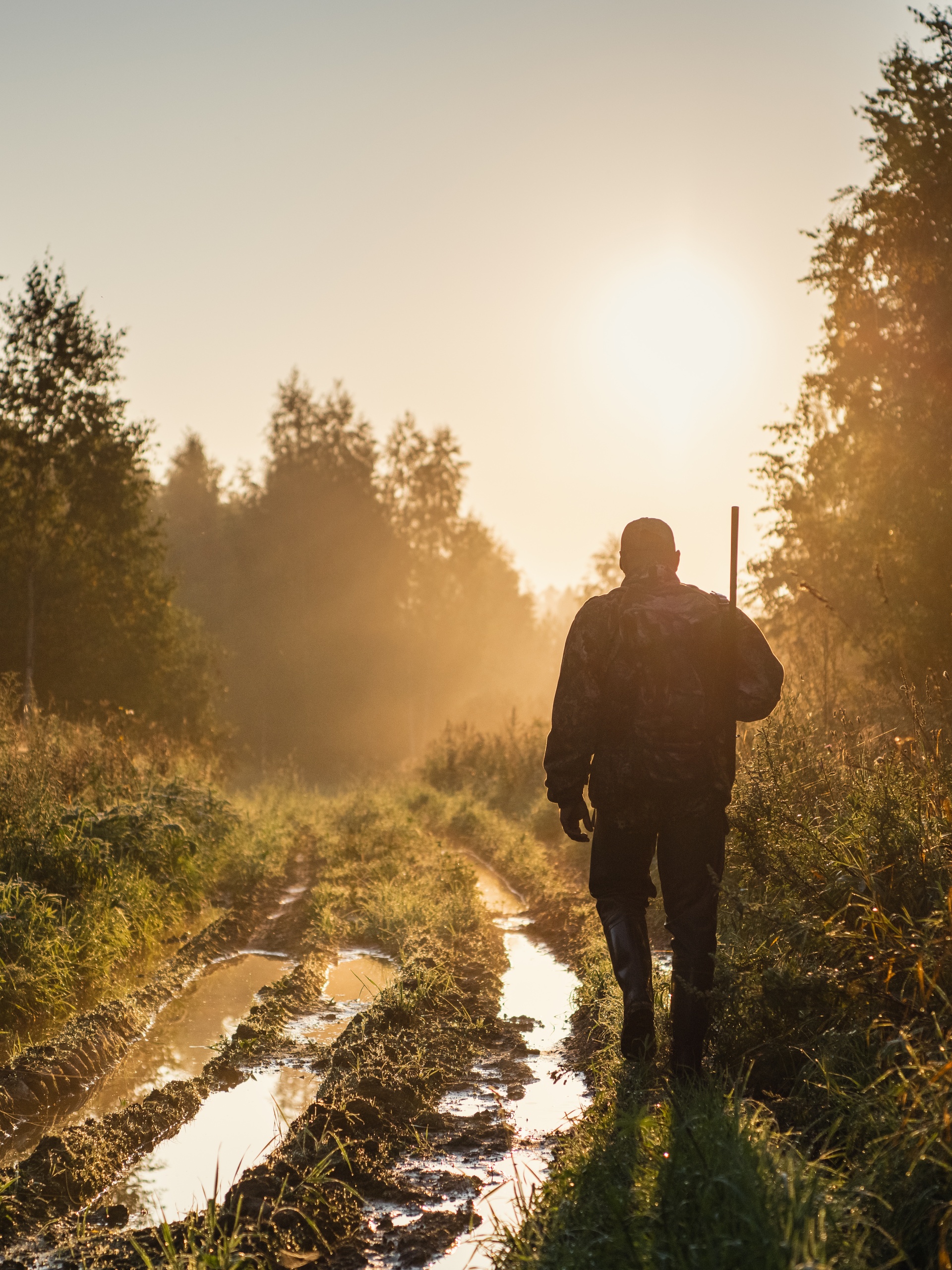 A hunter walking through a muddy field with a shotgun over his shoulder. The sun is setting in front of him.