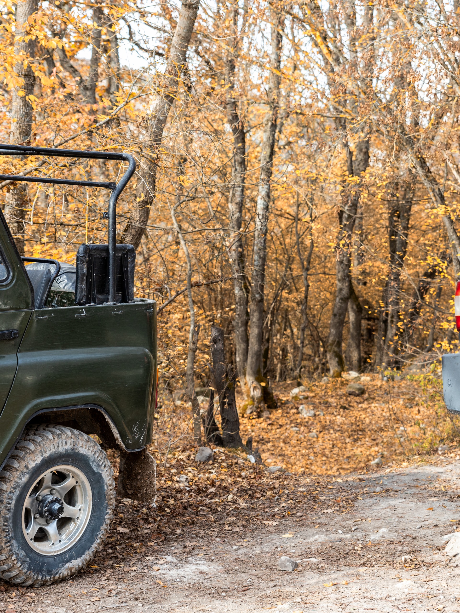 Two off-road vehicles sit on a dirt trail in a fall forest, with colorful fall leaves on trees in the background.
