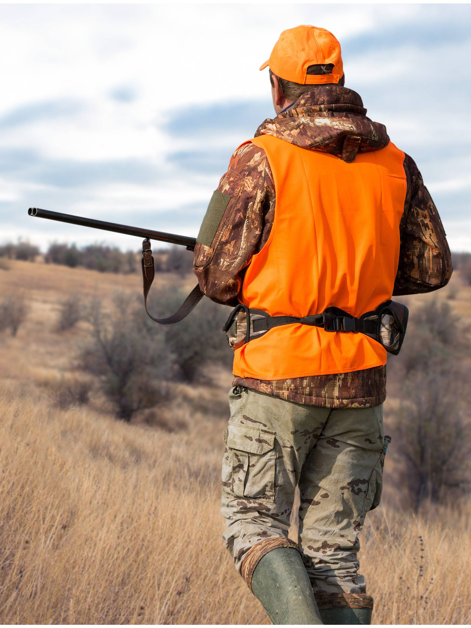A hunter in a blaze orange vest and camouflage clothing walks through a dry grassland field carrying a firearm over his shoulder.
