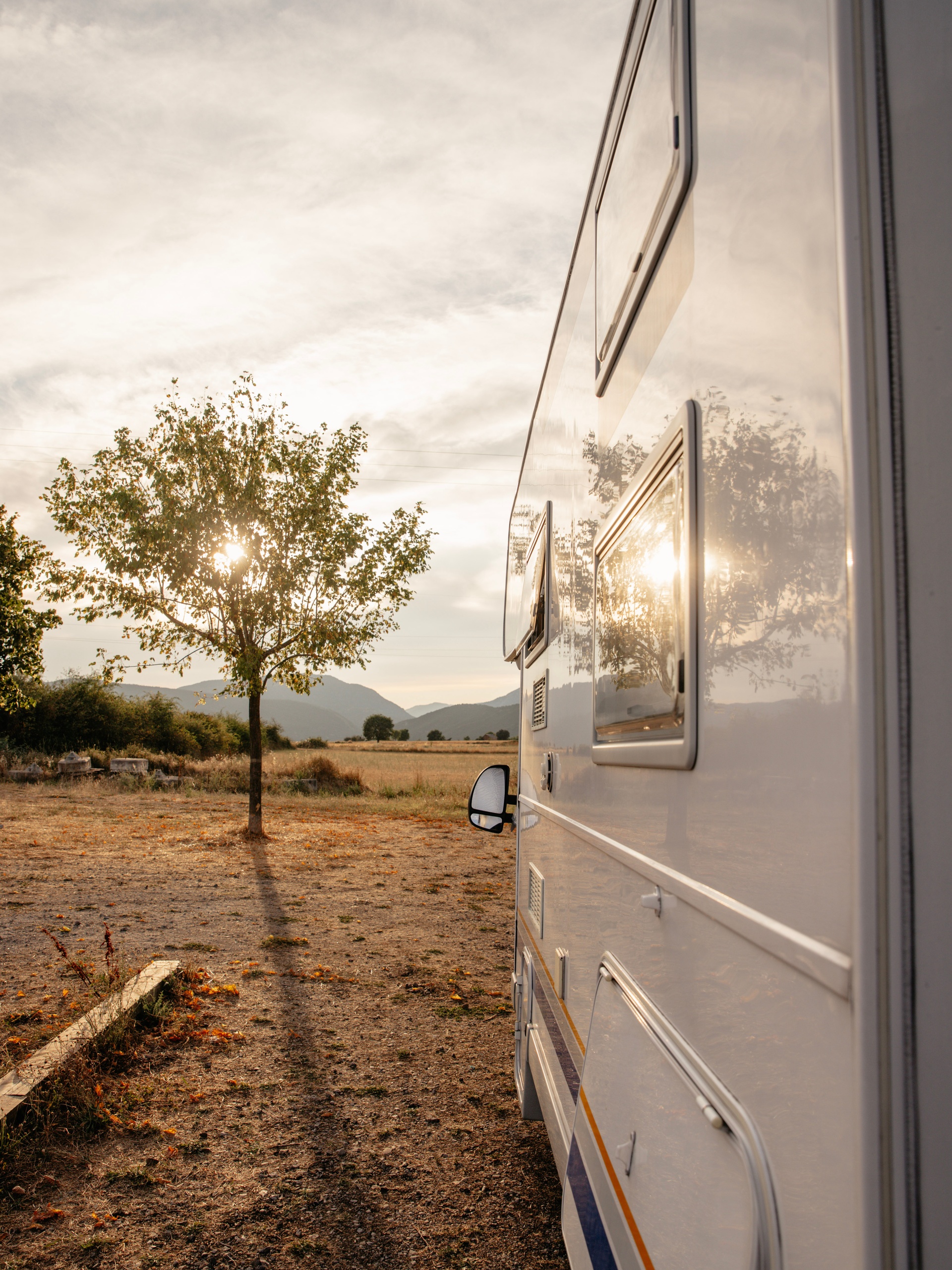 A white recreational vehicle parked beside a tree, with sunlight reflecting in its windows under a cloudy sky.