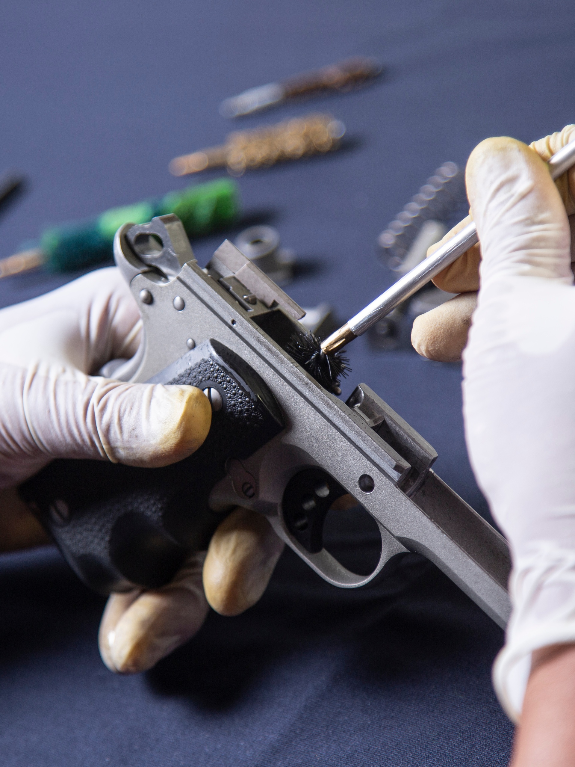 A person wearing white gloves cleaning a disassembled handgun with a metal brush on a dark work surface.