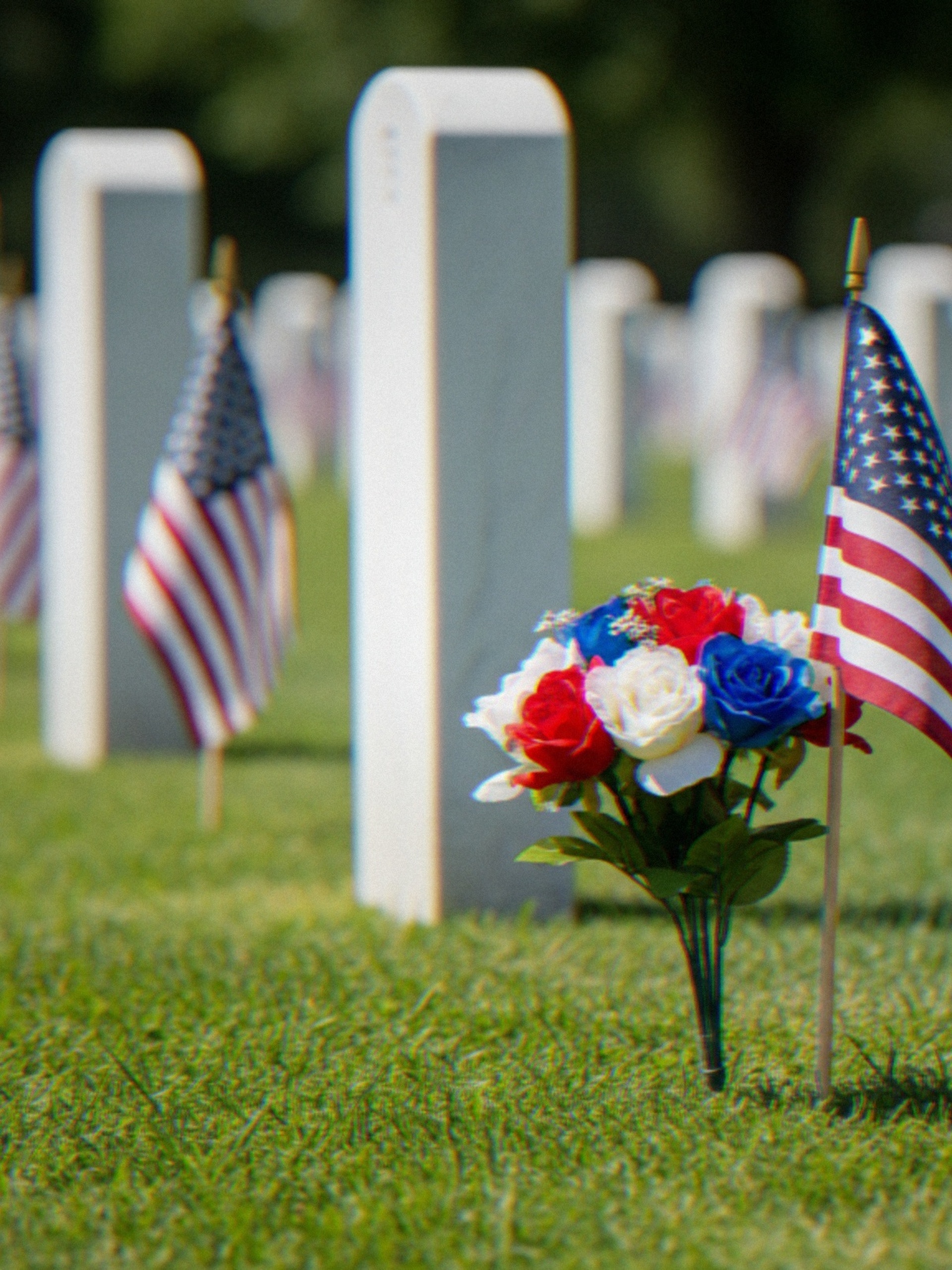 Rows of white gravestones with American flags and red, white, and blue flowers standing across a grassy cemetery.