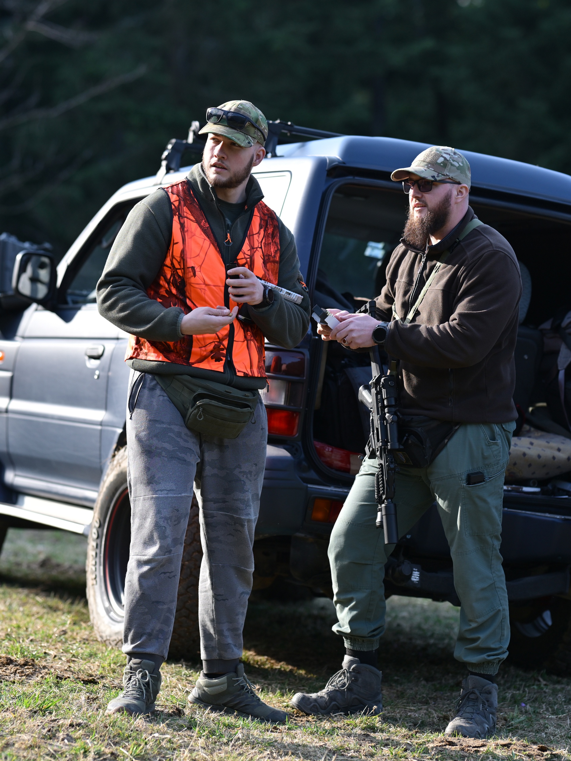 Two men on a hunting trip stand by a car with an open back door. One man wears an orange vest, and the other holds a firearm.